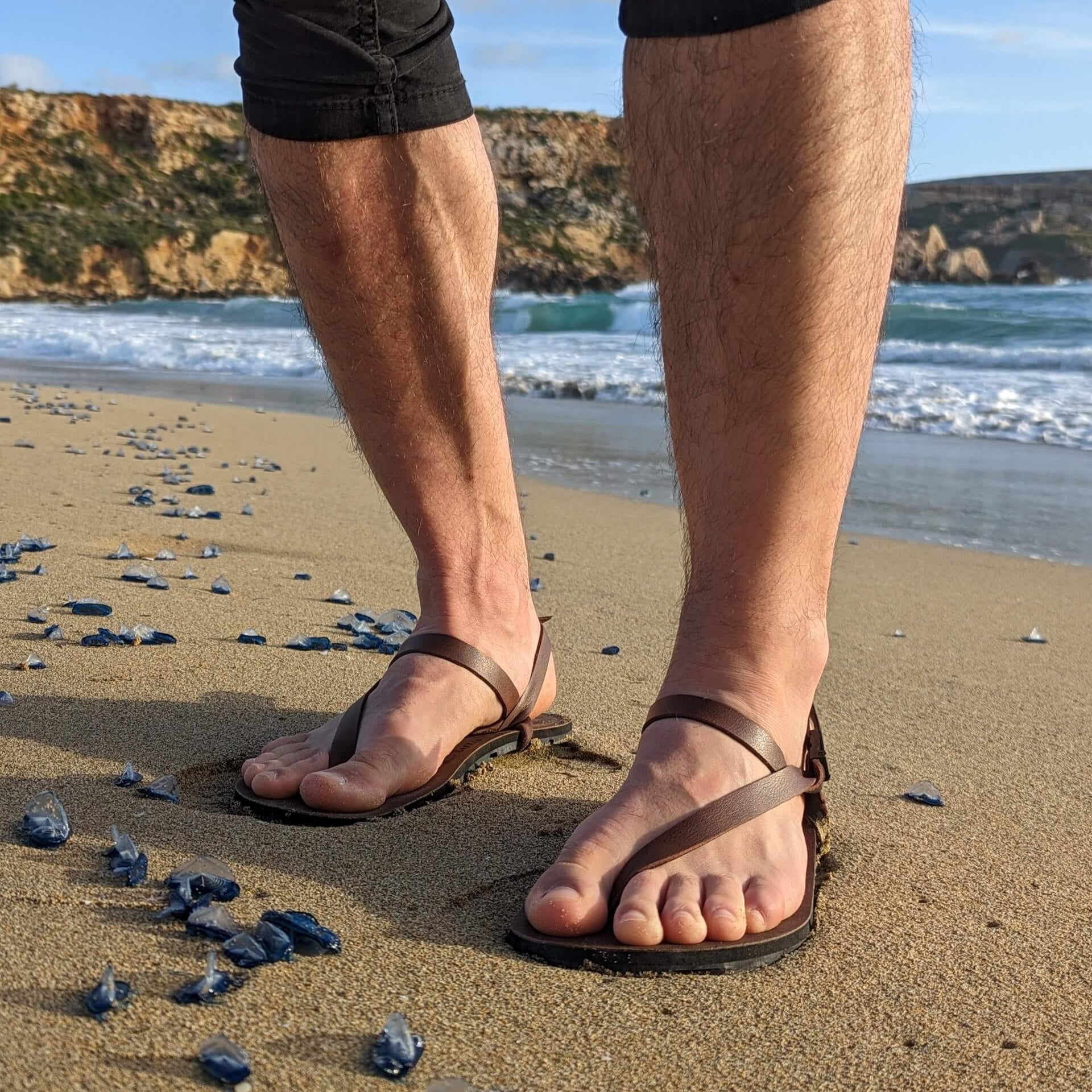 A man standing on the Shore line wearing ZuZu Groove barefoot sandals featuring vegan replaceable straps - perfect for everyday barefoot living. Sunset lighting and beautiful seashells.