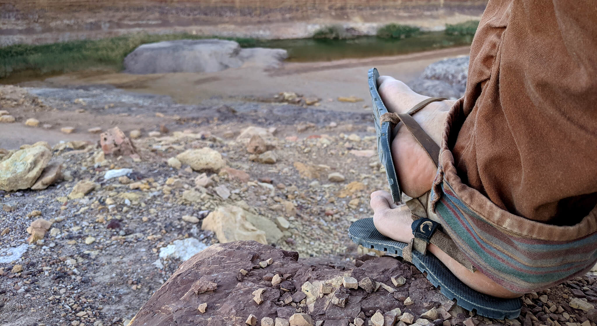 Feet wearing ZuZu Flex barefoot performance sandals with a breathable canvas footbed and replaceable recycled straps, designed for natural movement and comfort. Colorful sand background.